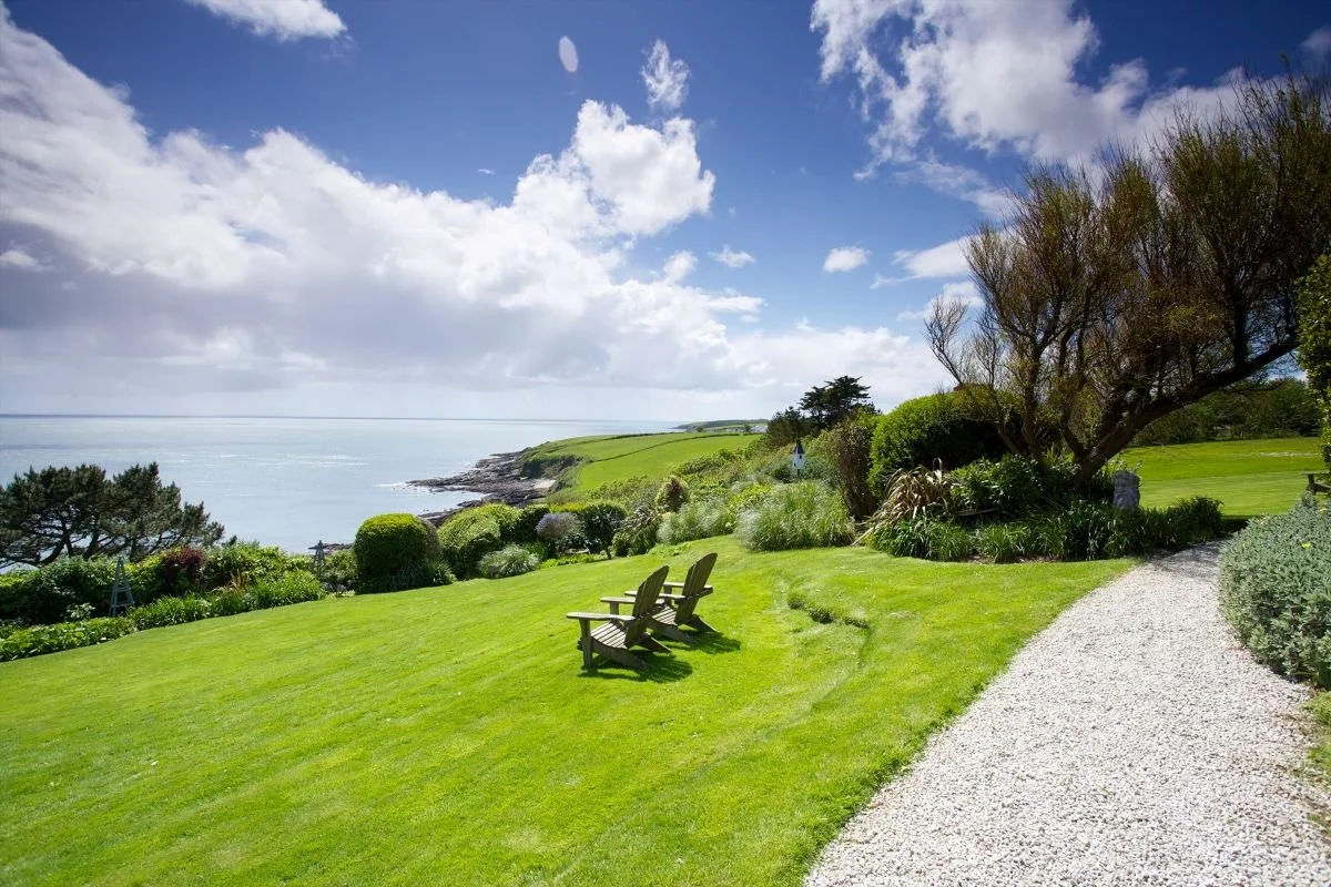 A scenic seaside landscape with green lawns, a gravel pathway, trees, bushes, and ocean in the background under a partly cloudy sky.