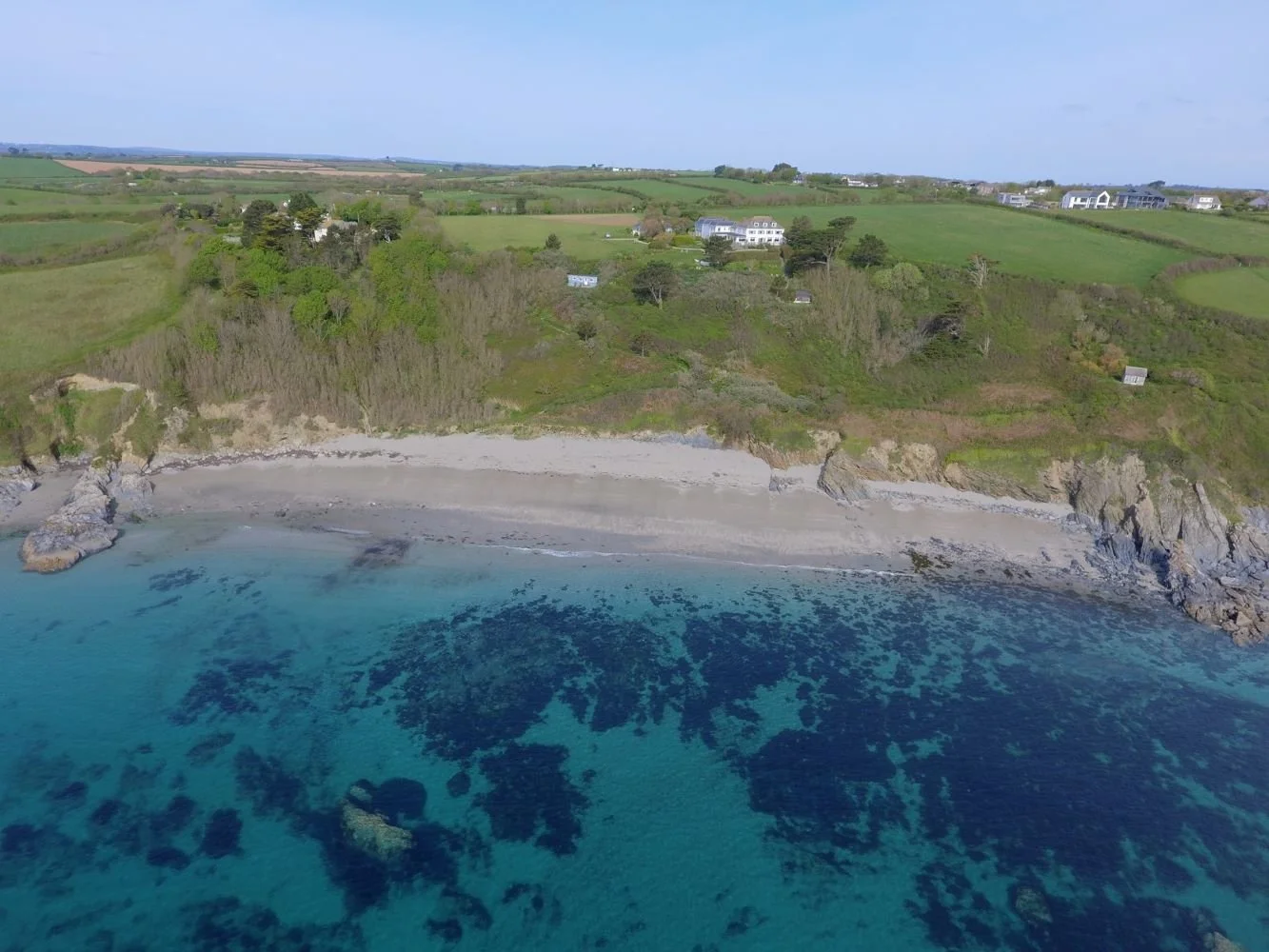 Aerial view of a coastal landscape with a sandy beach, turquoise water, rocky shoreline, and lush green trees and grass-covered hills with scattered houses and buildings in the background.