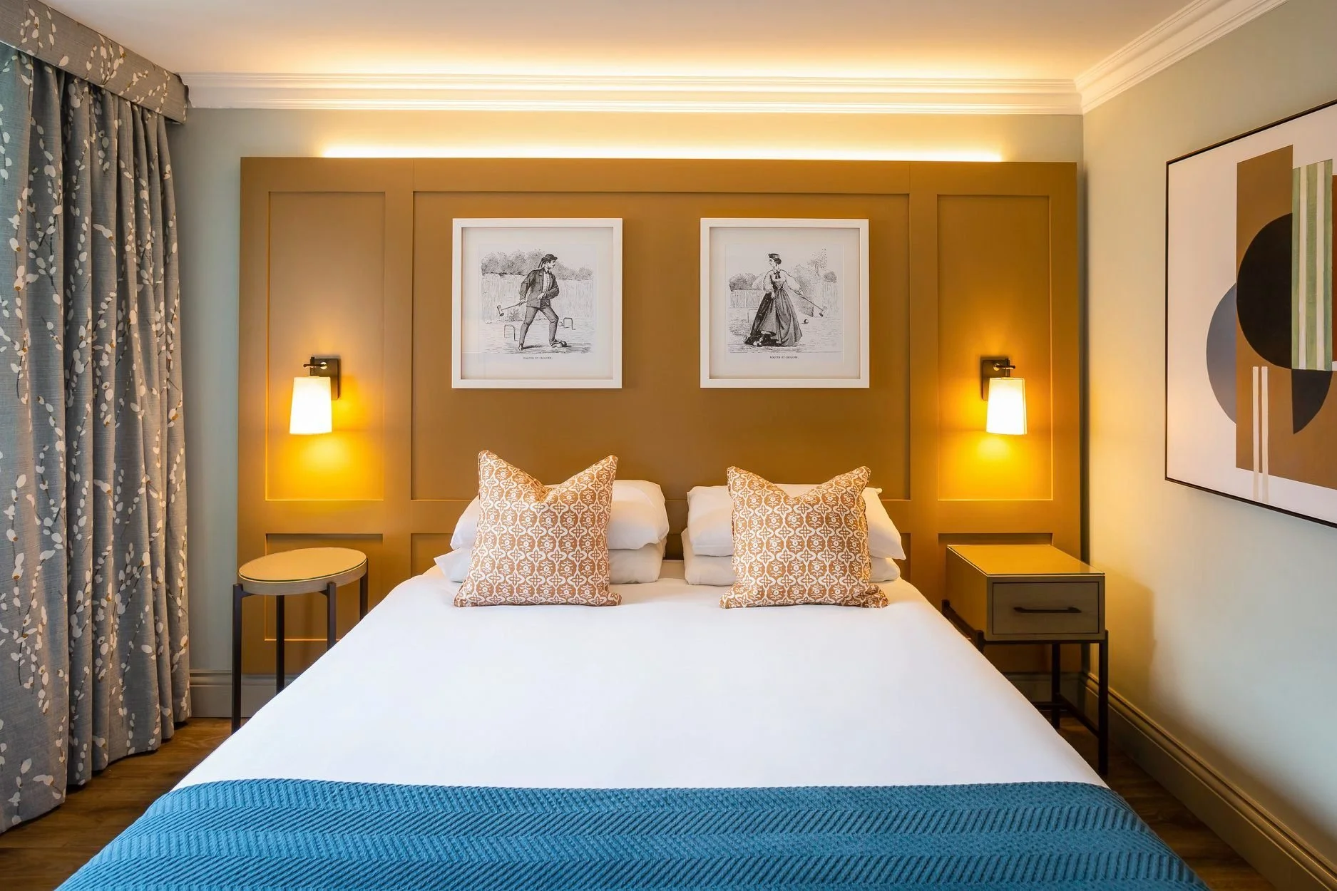 A hotel bedroom with a bed, two patterned pillows, and bedside tables with wall-mounted lamps. Framed artwork hangs above the headboard, and curtains with a leaf pattern are on the left side of the room.