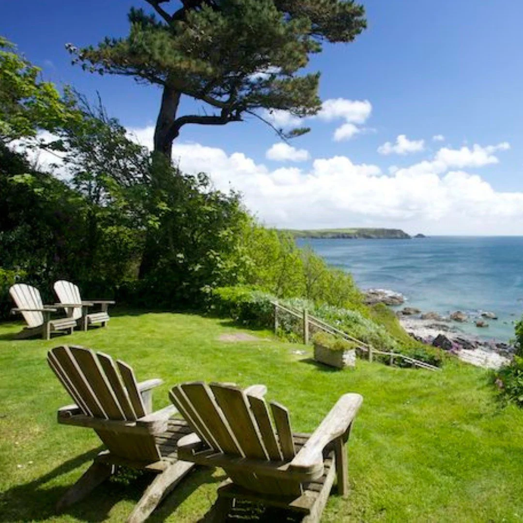 A peaceful coastal yard with green grass and Adirondack chairs, overlooking the ocean and rocky shoreline under a blue sky with scattered clouds.