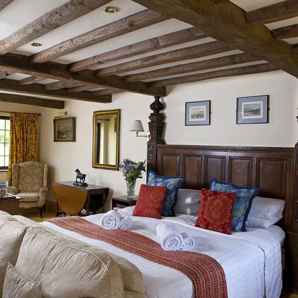A cozy bedroom with a wooden beamed ceiling, with neatly made bed featuring decorative pillows and rolled towels, a bedside table with flowers, and a vintage armchair by a window with floral curtains.