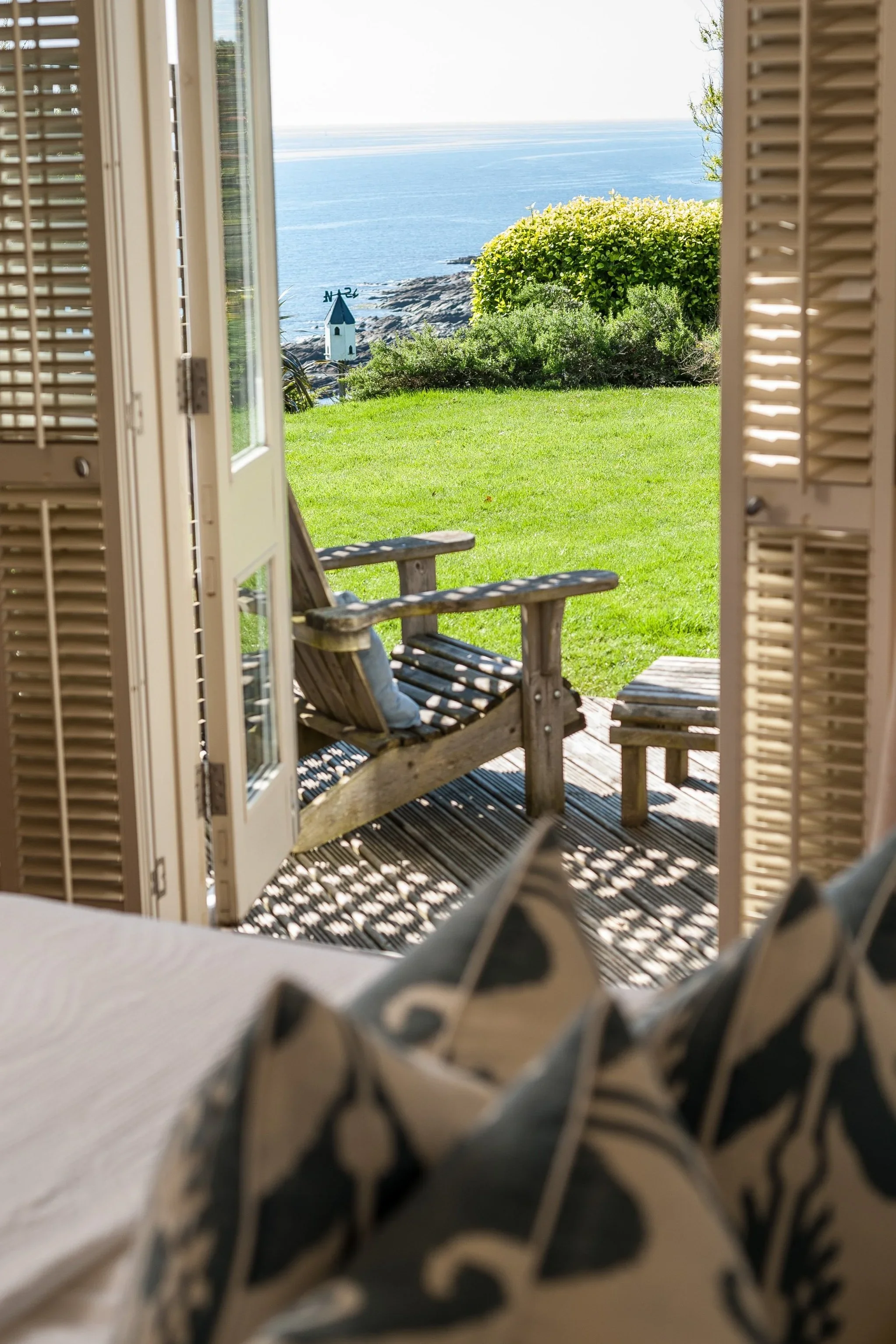 Open window with white shutters looking out onto a grassy patio area with Adirondack chairs, bushes, and a lighthouse by the ocean in the distance.