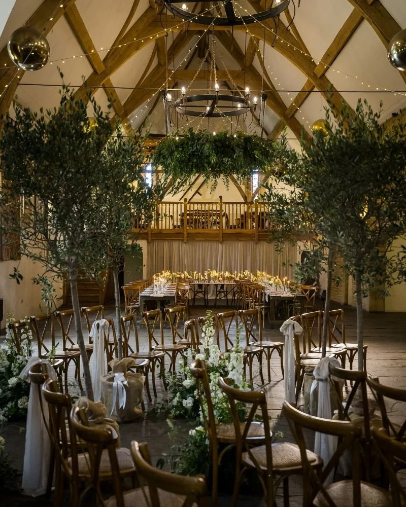 Wedding reception setup in a rustic barn with wooden chairs decorated with white ribbons, floral arrangements, greenery, and fairy lights hanging from the ceiling, creating a cozy and elegant atmosphere.
