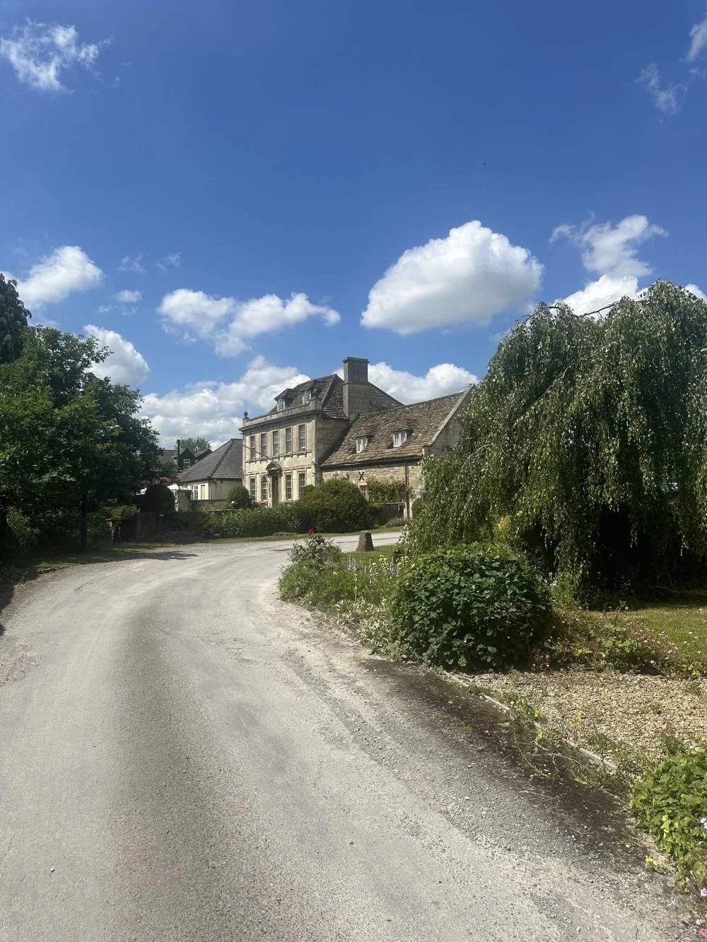 A country lane with a curve leading towards a large stone house with pitched roofs, surrounded by greenery under a blue sky with white clouds.
