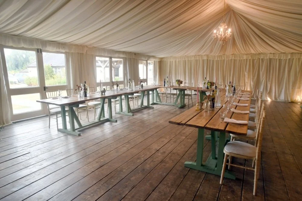 Empty event tent with long wooden tables arranged for a gathering, decorated with bottles and candles, and set with napkins and glassware, with large windows and a chandelier.