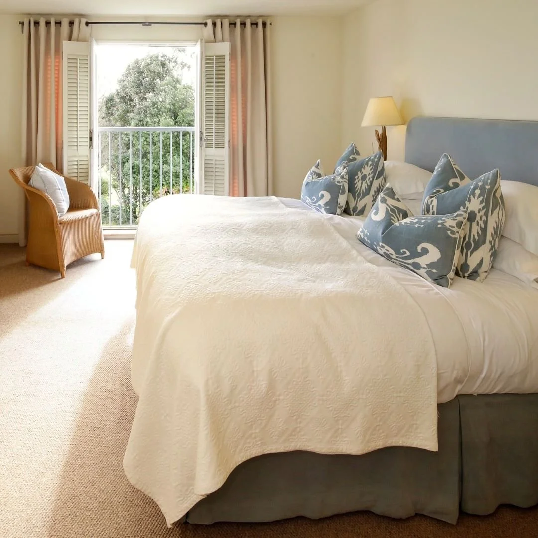 A cozy bedroom with a large bed, multiple pillows, a bedside lamp, a wicker chair with a cushion, and an open balcony door revealing greenery outside.