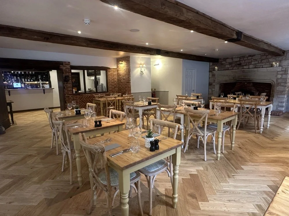 Empty restaurant dining area with wooden tables, chairs, and a fireplace, set for service with water glasses, cutlery, and small decor.