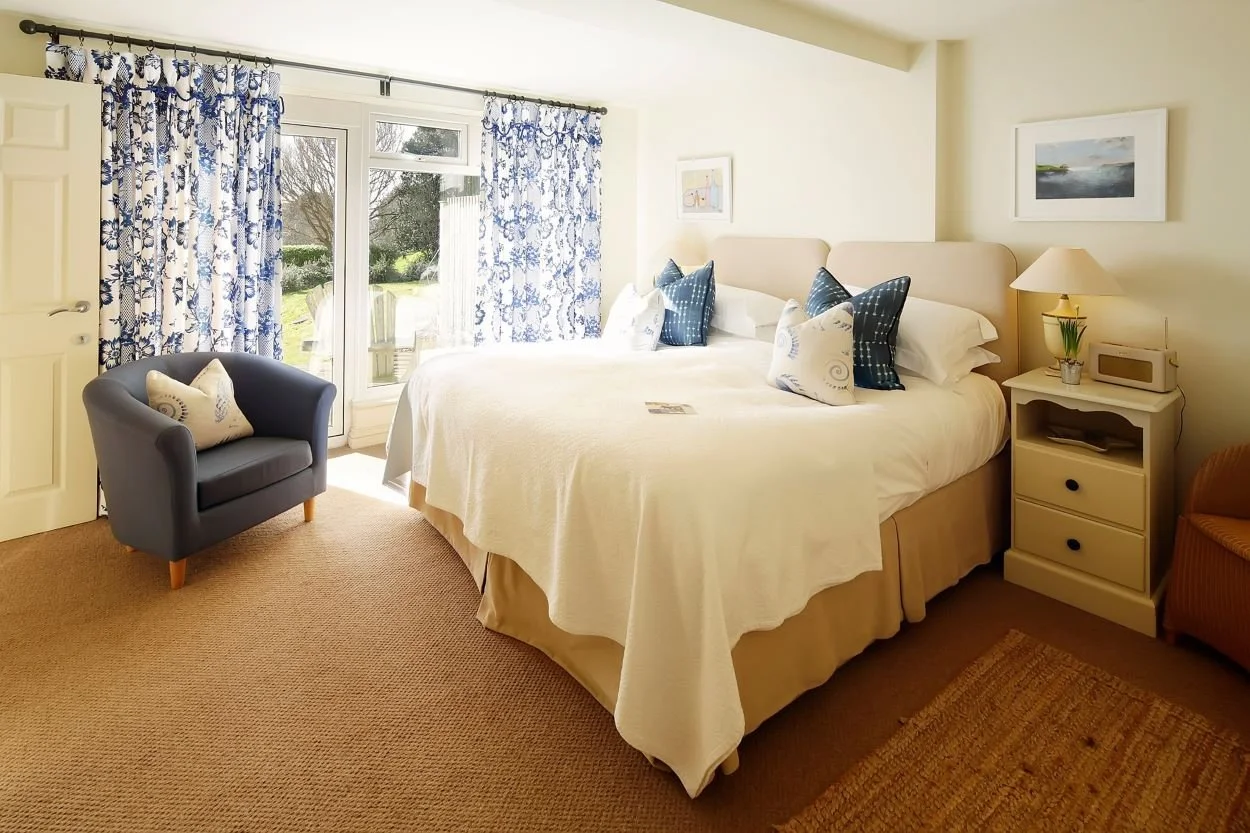 Bright hotel bedroom with a large bed, nightstand, armchair, and sliding glass door with blue patterned curtains.
