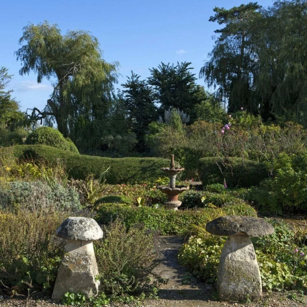 A garden scene with stone mushroom-shaped sculptures at the entrance, a small stone fountain in the background, lush green bushes, trees, and a clear blue sky.