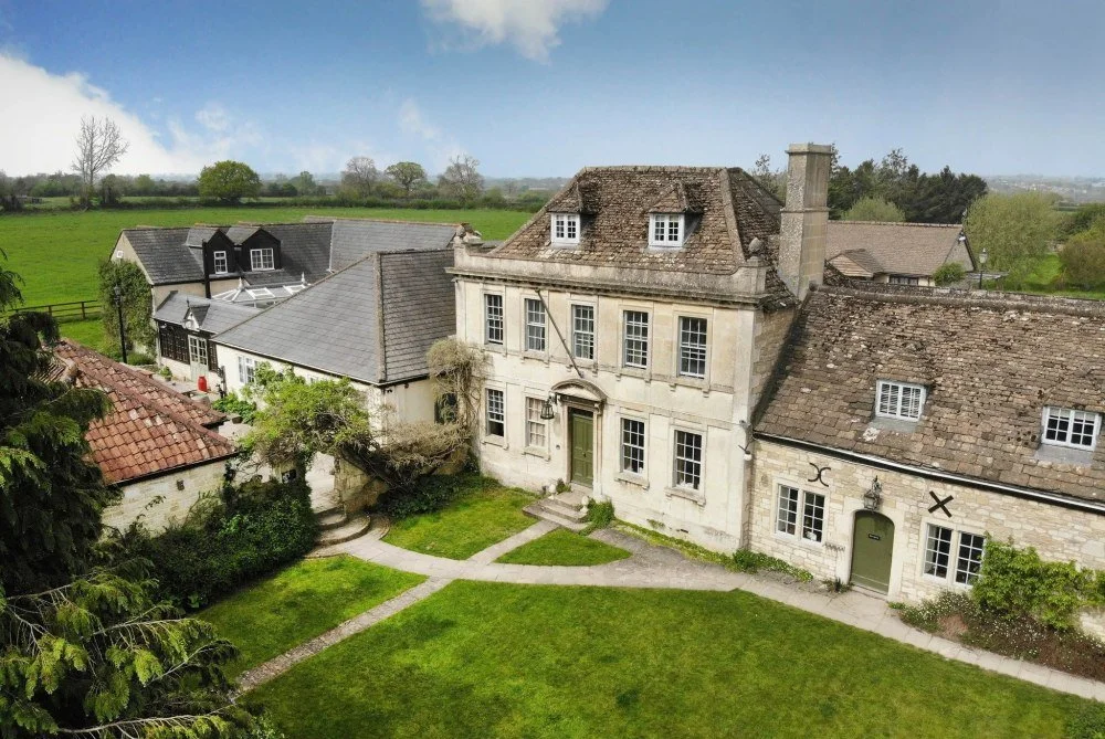 A historic stone house with multiple levels, surrounded by a well-maintained lawn and pathways, in a rural setting with other buildings and green fields in the background.