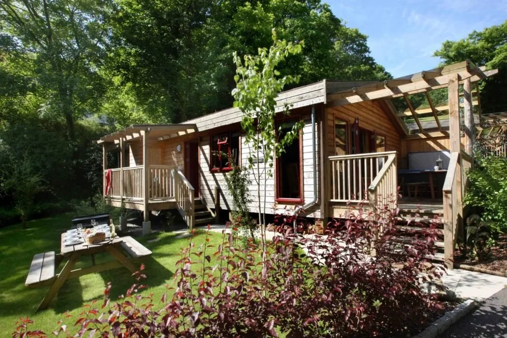A small wooden house with a deck, surrounded by greenery and trees, with a picnic table in the yard.