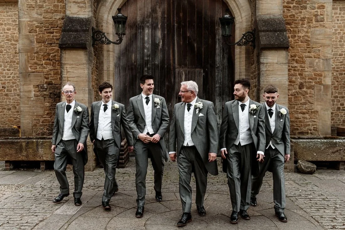 Group of men in gray suits walking and smiling outside a stone building with a large wooden door.