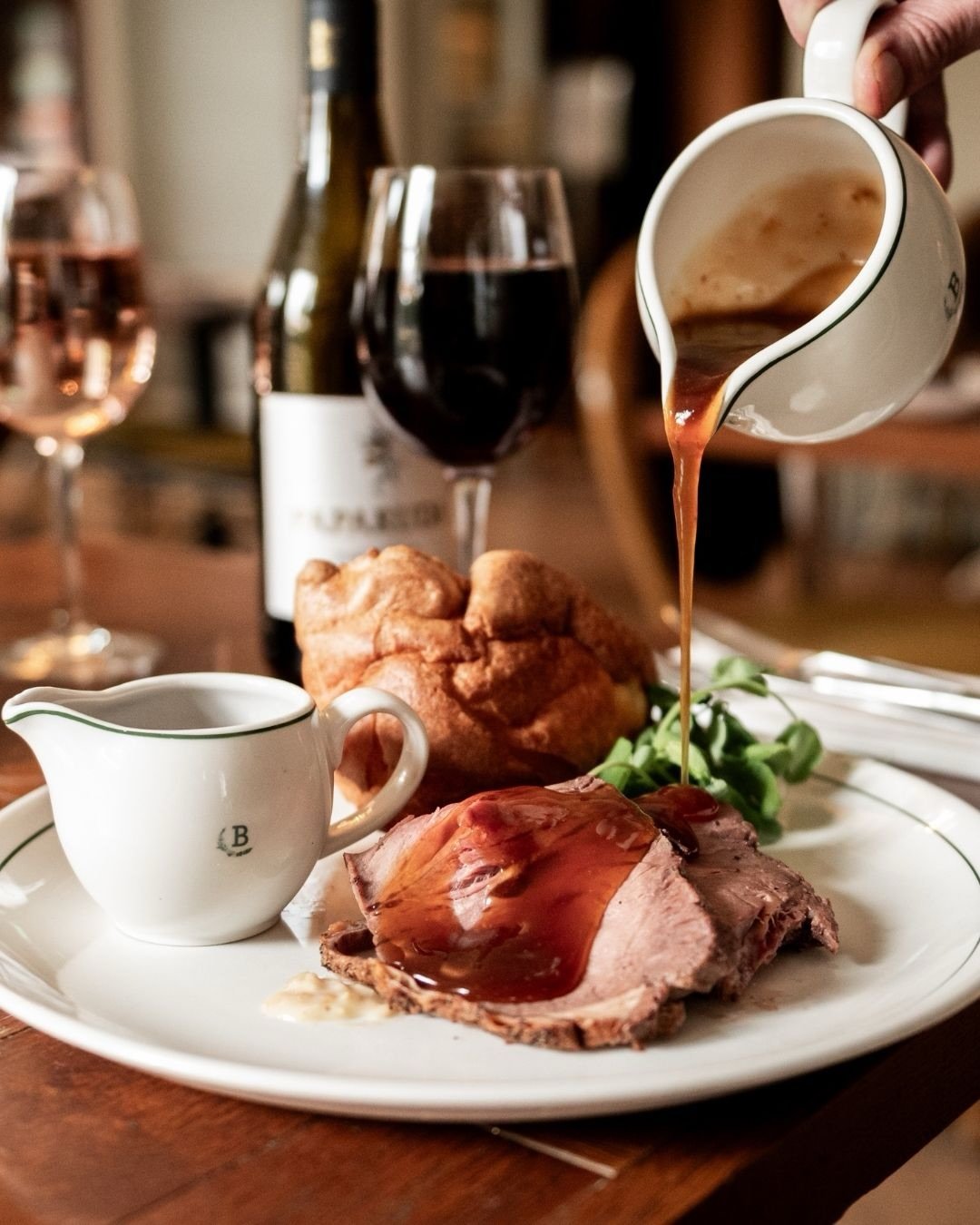 A plate with roast beef, gravy, green herbs, and a Yorkshire pudding. A small pitcher of gravy is on the plate. In the background, glasses of red wine, a bottle, and a person's hand pouring gravy from a white pitcher.