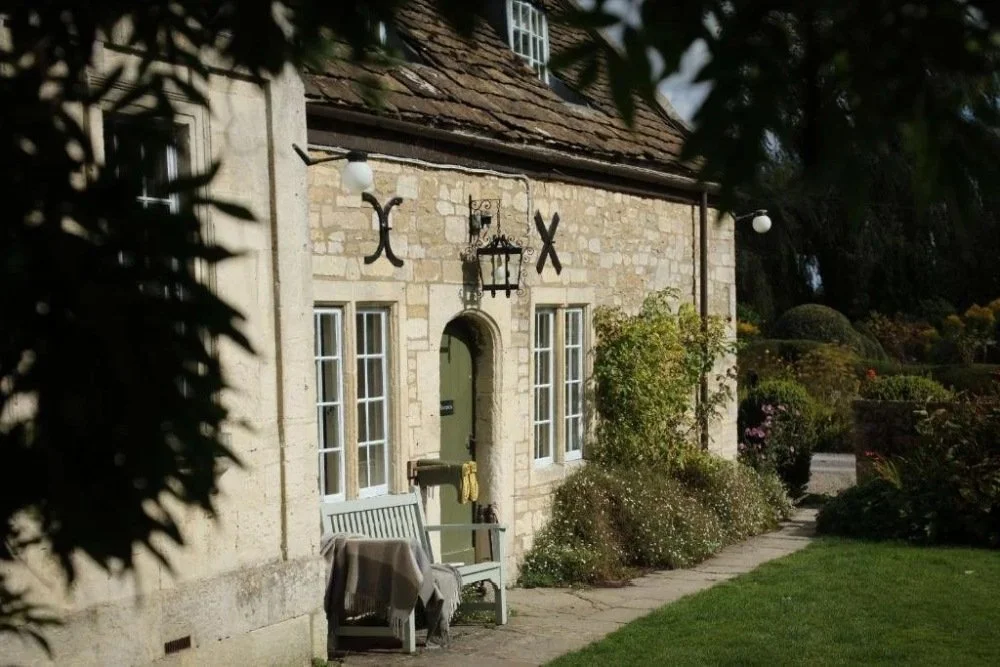 A stone cottage with a shingled roof, surrounded by bushes and a grassy garden, with a bench and a table outside near the door.