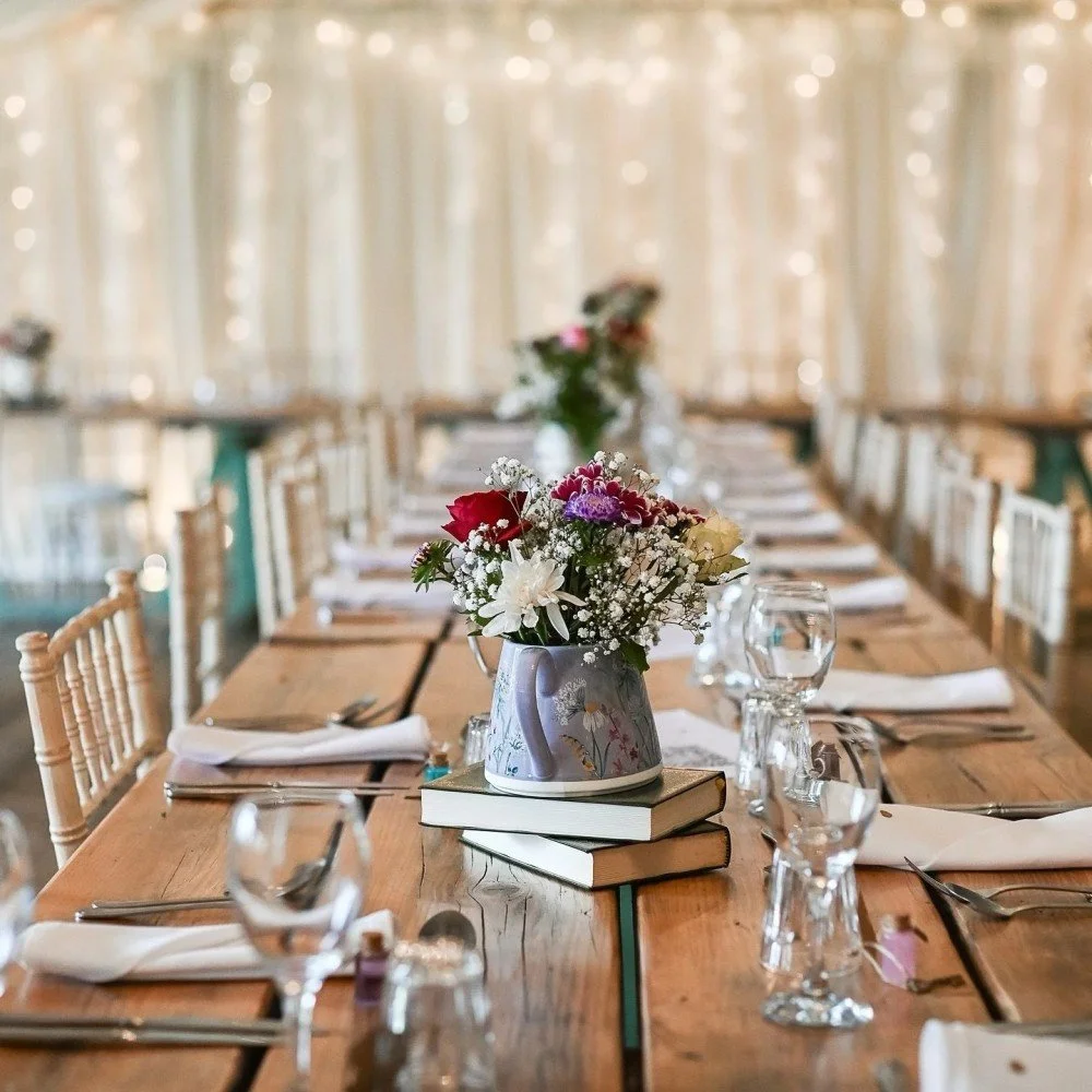 Long wooden dining table set with white napkins, silverware, clear glass water glasses, and a centerpiece of flowers in a watering can on top of stacked books.
