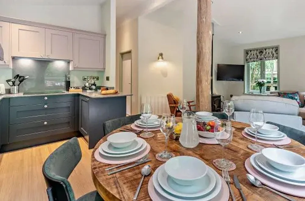 A dining table set with plates, bowls, wine glasses, a creamer, and a fruit bowl. In the background, a kitchen with pastel-colored cabinets and a living room with a TV and a window.