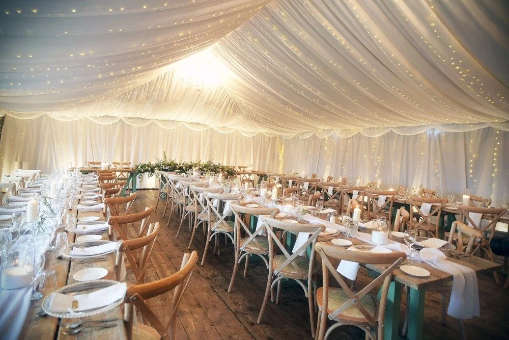Elegant event tent decorated with white drapes and string lights, set with long wooden tables and chairs for dining.