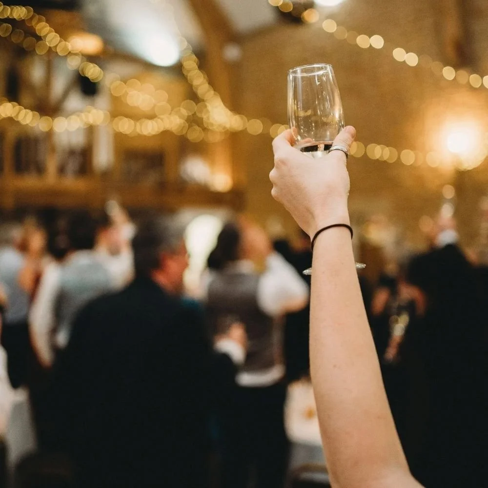 A person holding a glass of champagne at a crowded celebration or event, with warm string lights and blurred guests in the background.
