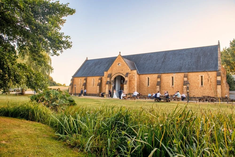 A historic brick church with a large sloped roof, surrounded by greenery and people seated outside on benches, during daytime.