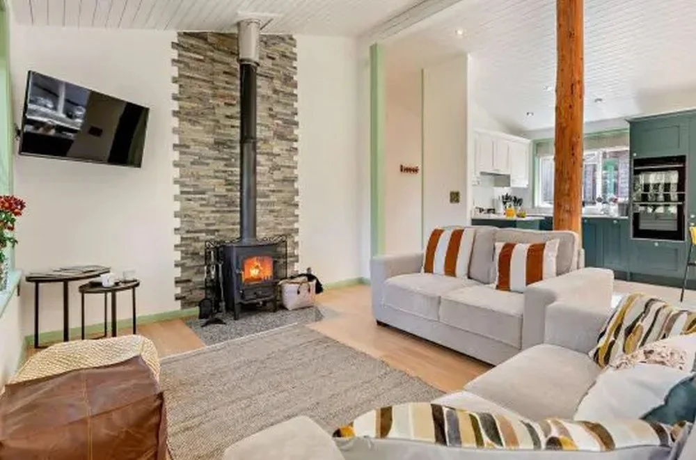 Living room with a fireplace, a wall-mounted TV, two sofas with striped cushions, a textured stone chimney, and an open kitchen in the background.