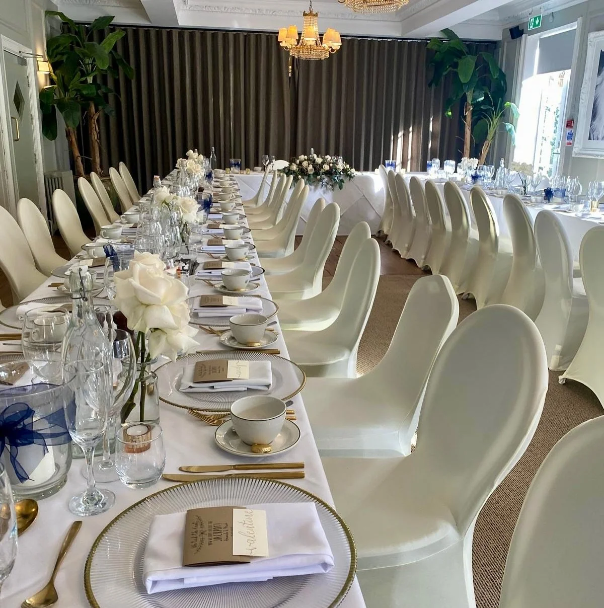 Elegant dining table set for a wedding or special event with white chairs, gold accents, white flowers, and formal place settings, in a bright room with curtains and large plants.