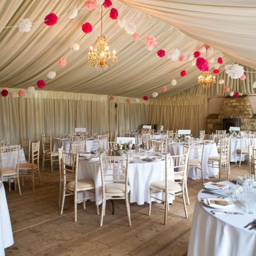 Decorated banquet hall with cream-colored draped ceiling, pink, white, and red hanging paper flowers, chandeliers, and round tables set for a formal event.