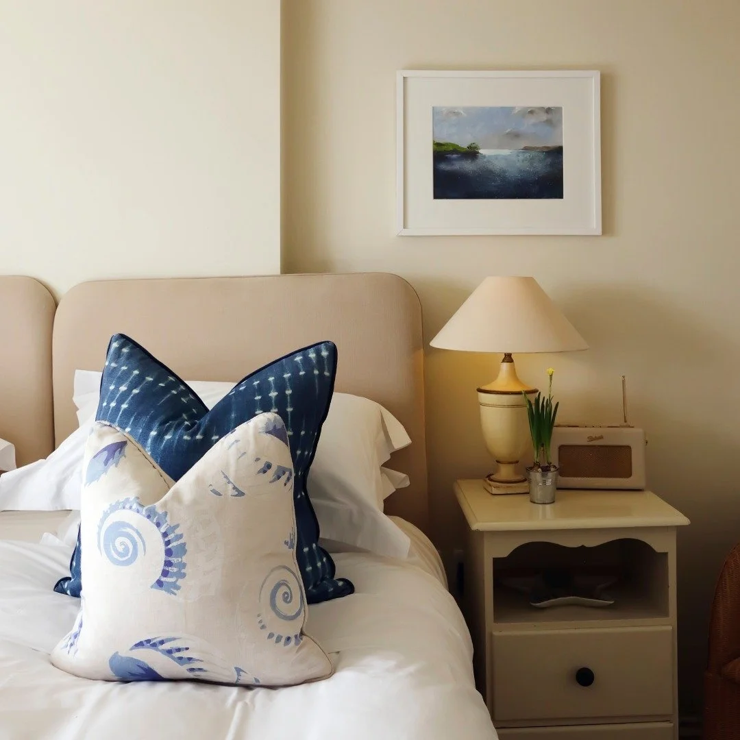 A bedroom with a beige headboard, ornate pillows in blue and white, a white nightstand with a lamp, a small potted plant, and a framed landscape painting on the wall.
