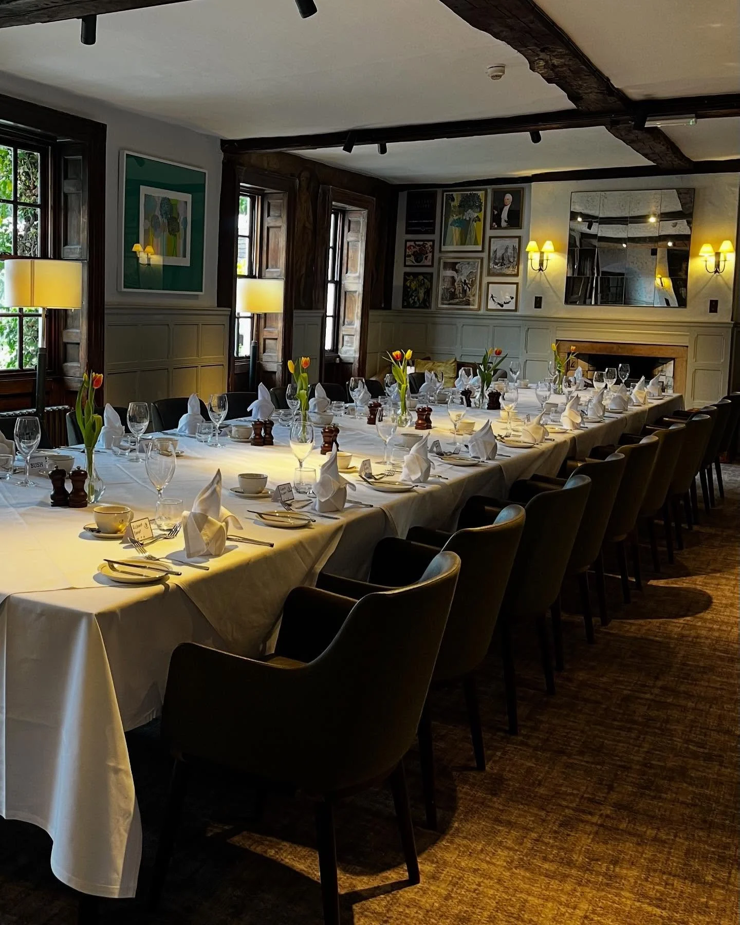 A long dining table set for a meal with white tablecloth, wine glasses, folded cloth napkins, small flower arrangements, and salt and pepper shakers in a warmly lit room with artwork on the walls and wooden window frames.