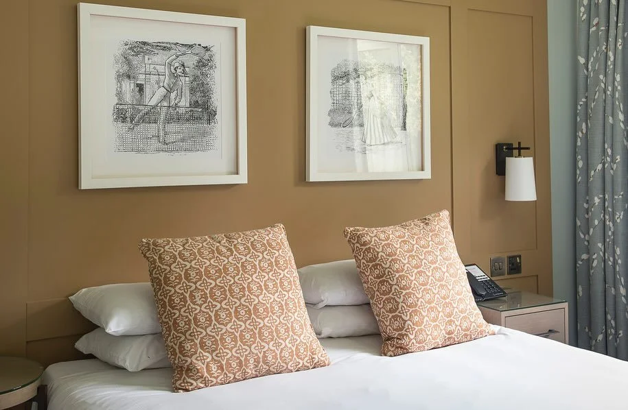 Hotel bed with two large patterned pillows, white sheets, and side table with phone and lamp in a neutral-colored room.