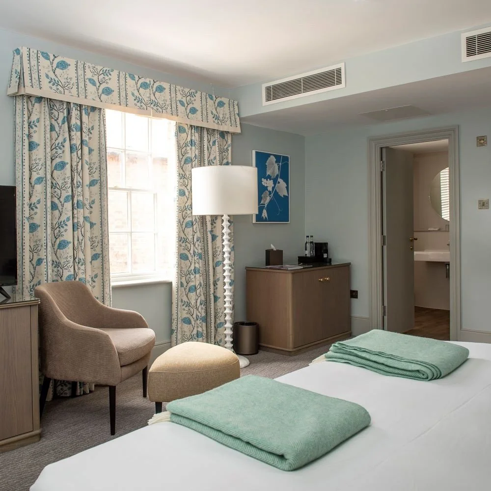 A hotel room with a bed covered with two green blankets, a chair with an ottoman, a window with patterned curtain, a floor lamp, a cabinet with water bottles and a coffee maker, and a doorway leading to a bathroom.