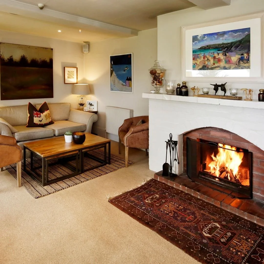 Living room with beige sofa, armchair, wooden coffee table, fireplace with a roaring fire, wall art, and decorative items.
