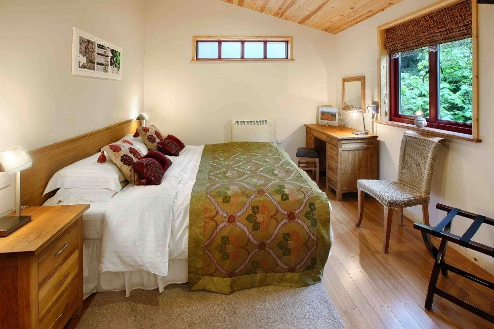 Cozy bedroom with wooden furniture, a bed with decorative pillows, a nightstand with a lamp, a small desk with a mirror, and a chair near a window showing greenery outside.