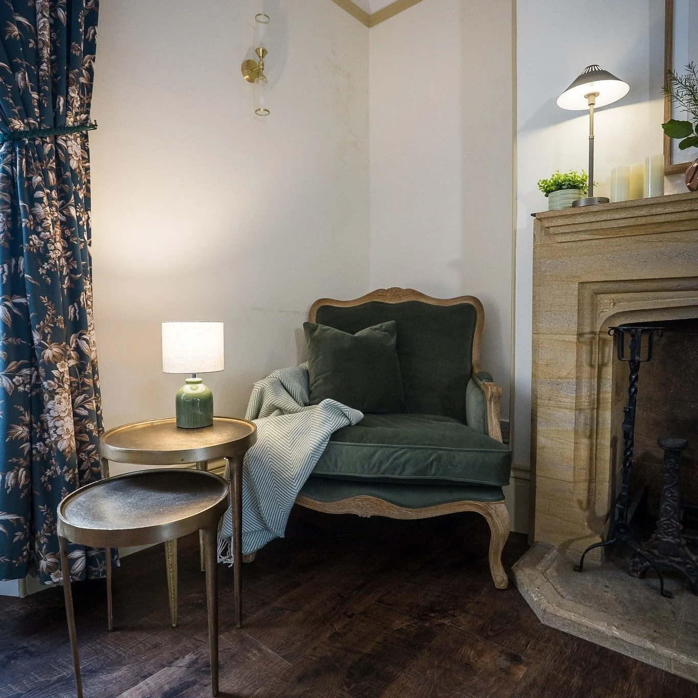 A cozy corner with a vintage green velvet armchair, a small round side table with a green ceramic lamp, a patterned curtain, and a fireplace with a pine mantel decorated with candles and a plant.
