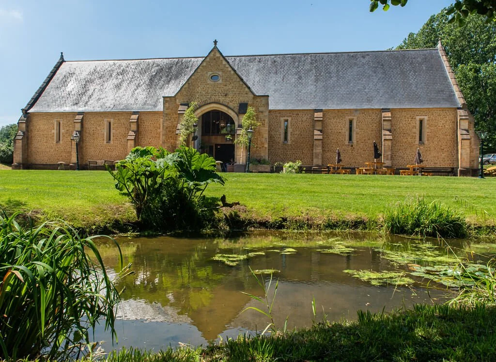 An old stone church with a pitched roof, situated on a grassy bank beside a pond with lily pads, under a clear blue sky.