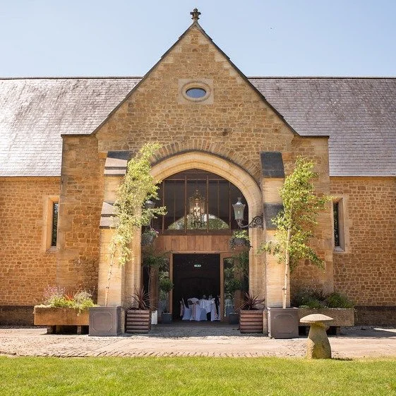 Front view of a historic stone church or chapel with an arched entrance, two small trees on either side, and outdoor tables inside, set up for an event, with clear blue sky.