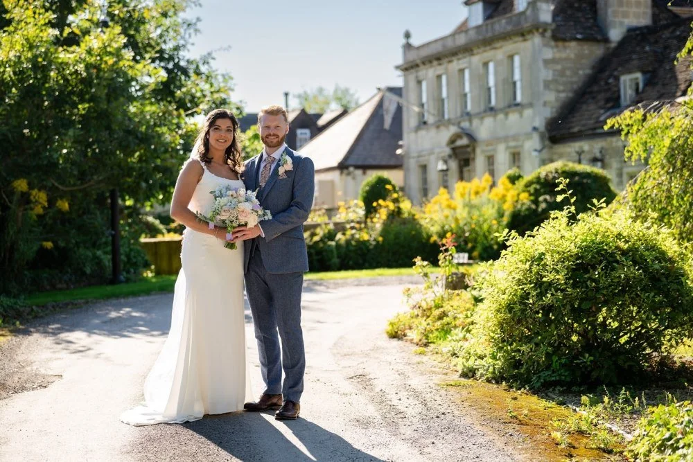 A bride and groom standing outside on a sunny day, posing for a wedding photo. The bride is holding a bouquet of flowers, and both are smiling, dressed in wedding attire, with a historic house and lush greenery in the background.