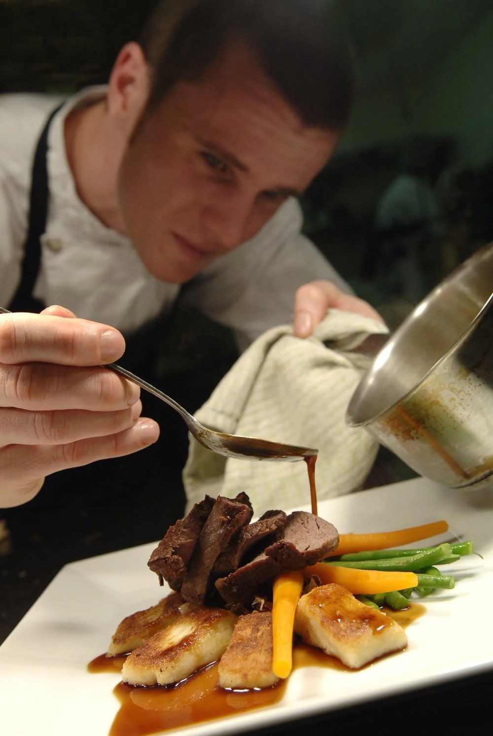 Chef pouring sauce over a plated dish of beef, seared fish, and vegetables.