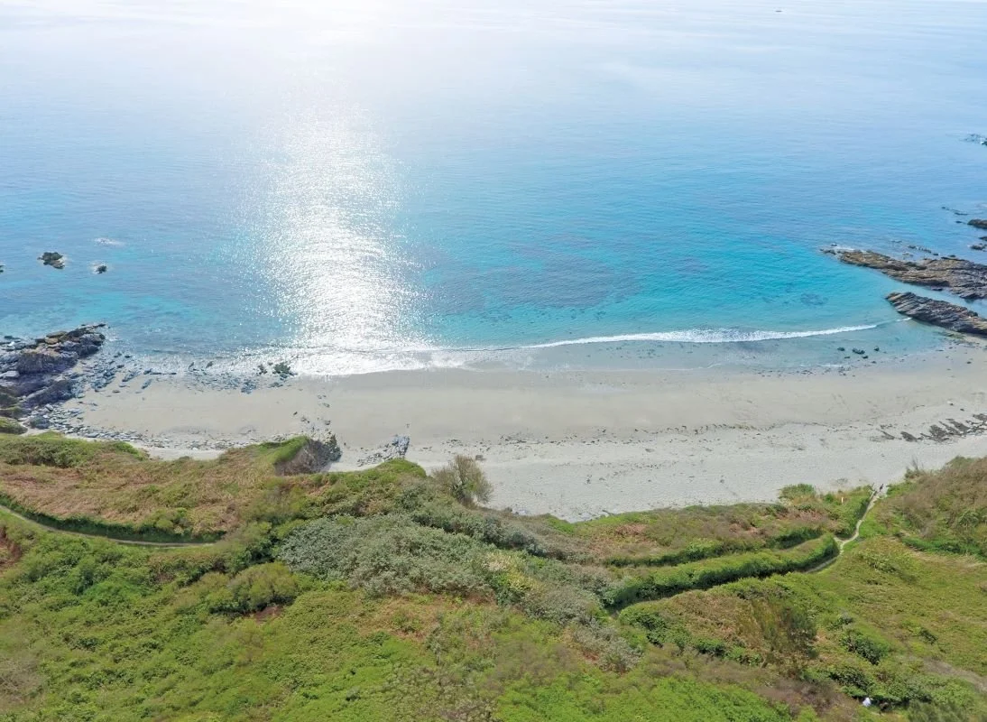 Aerial view of a beach with rocks, white sand, and clear blue water, bordered by green vegetation and hills.