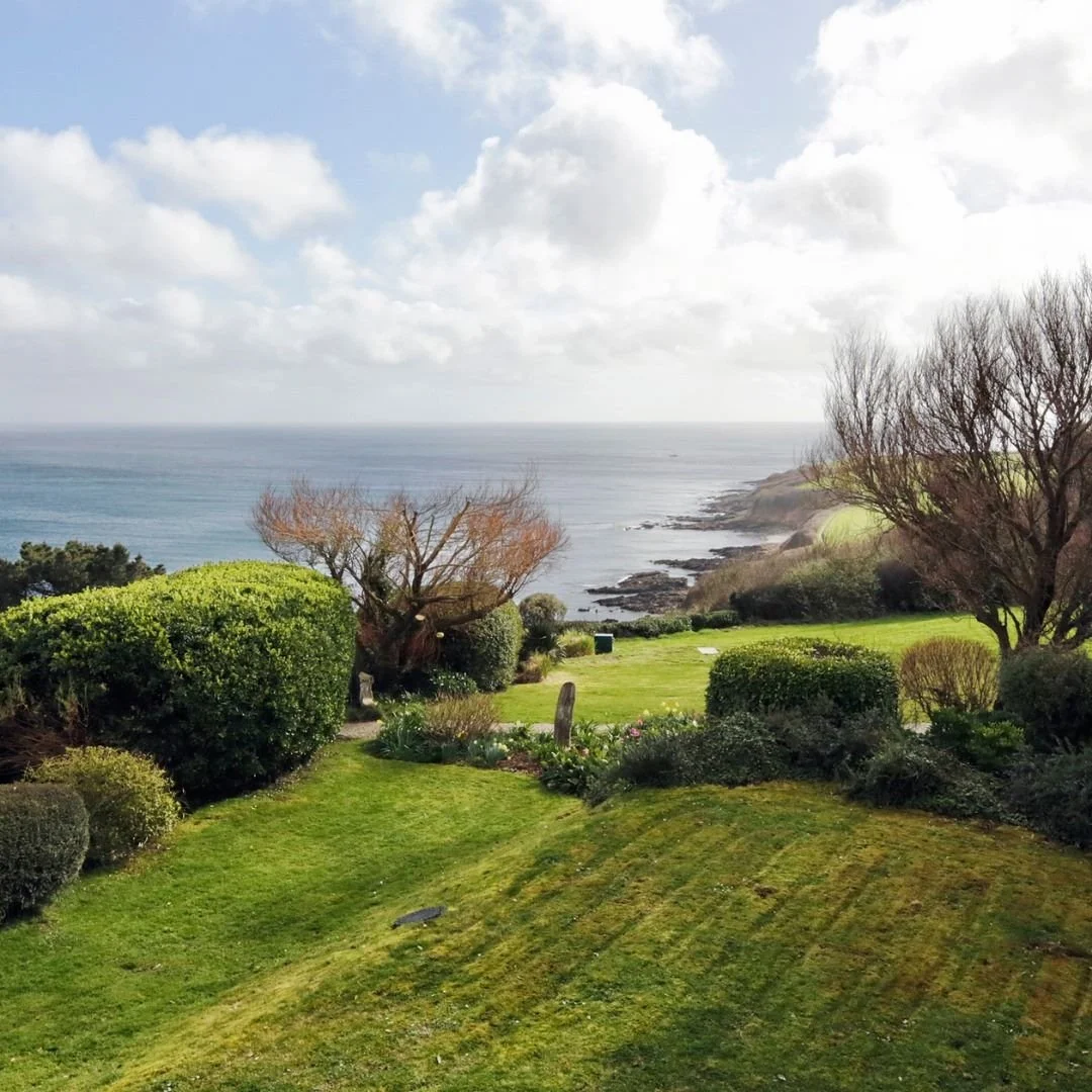 Scenic view of a lush green garden leading to the ocean with trees, bushes, and a rocky shoreline under a partly cloudy sky.