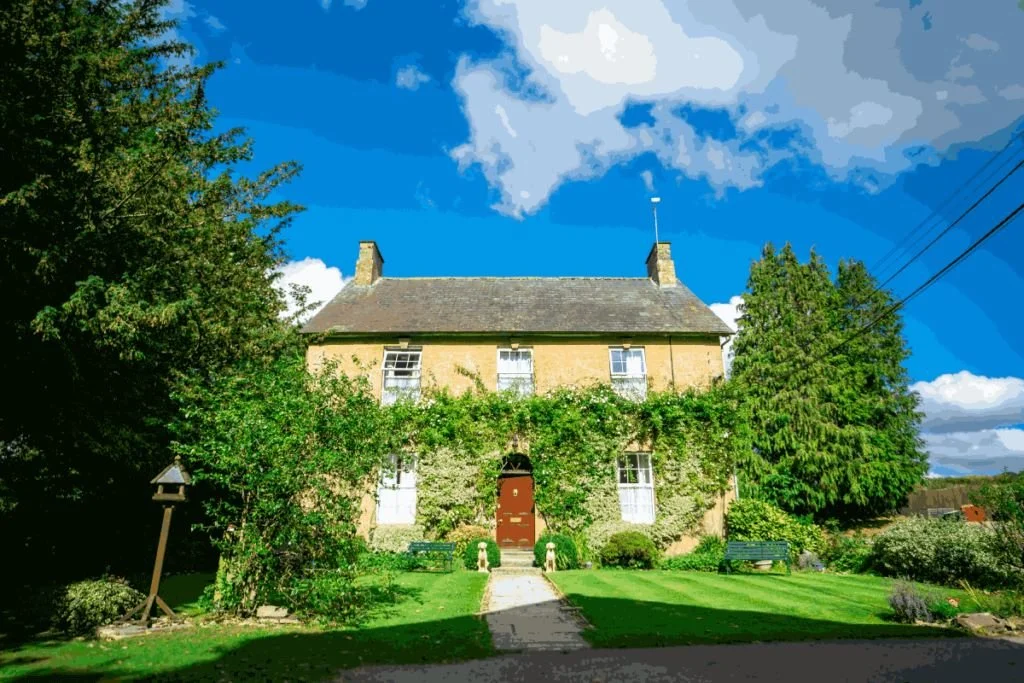A yellow two-story house with a steep roof, chimney stacks, and a red door, surrounded by lush green trees and bushes, under a partly cloudy blue sky.