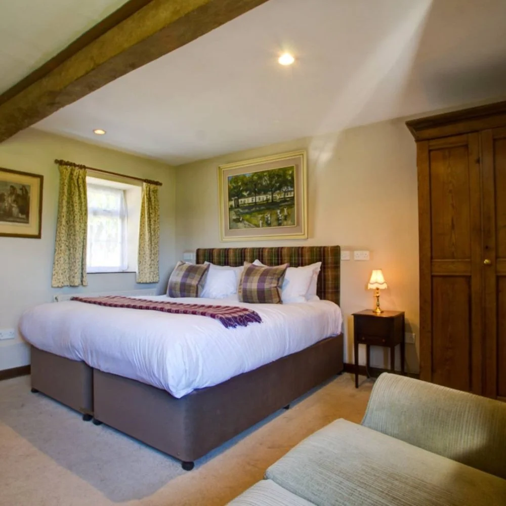 A cozy bedroom with a large bed featuring a plaid headboard, white bedding, and multiple pillows, situated next to a nightstand with a small lamp. There is a window with patterned curtains and a framed painting on the wall. A green armchair is partia