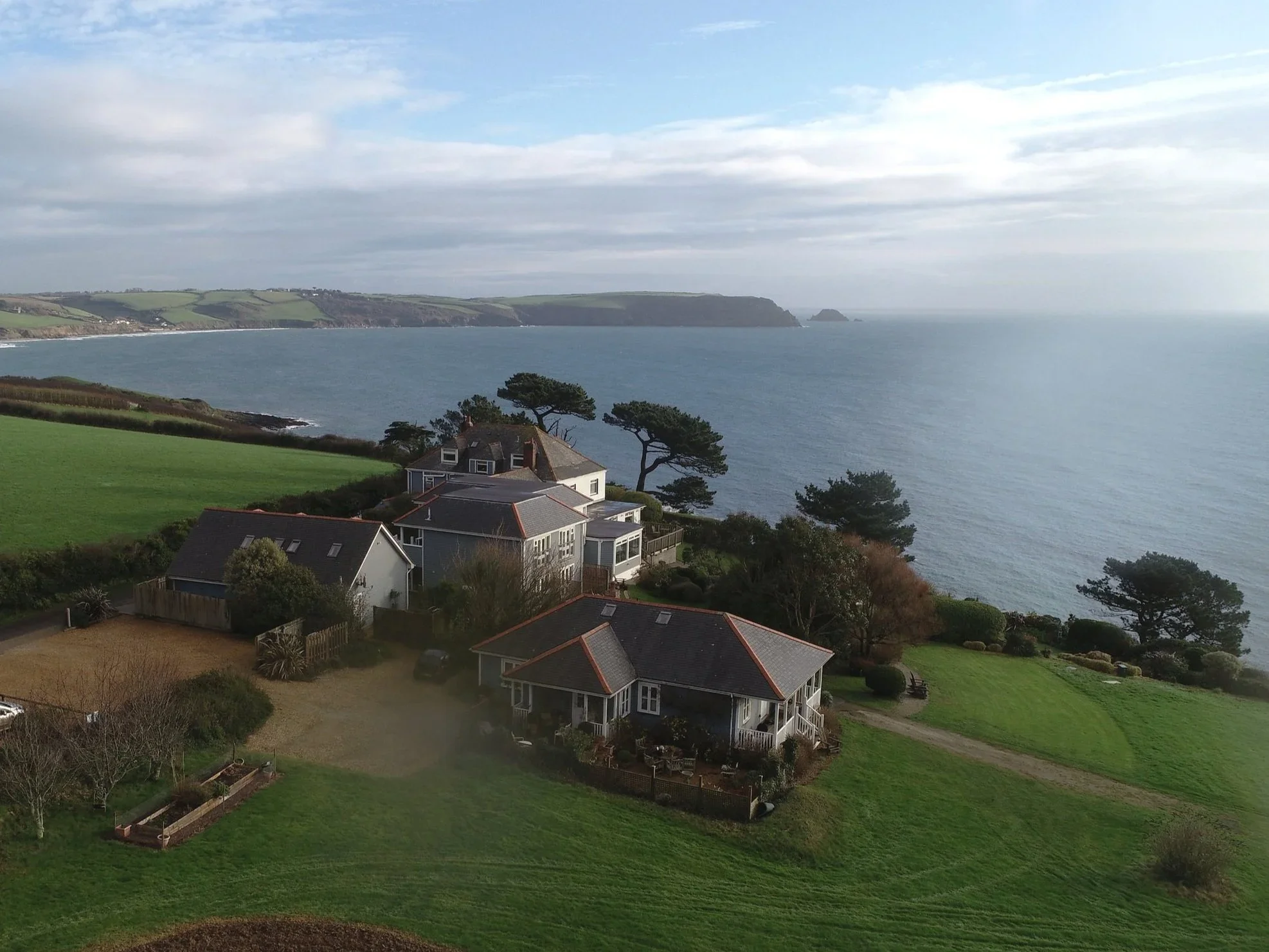 A coastal landscape with houses and trees in the foreground, a body of water beyond, and cliffs in the distance under a partly cloudy sky.