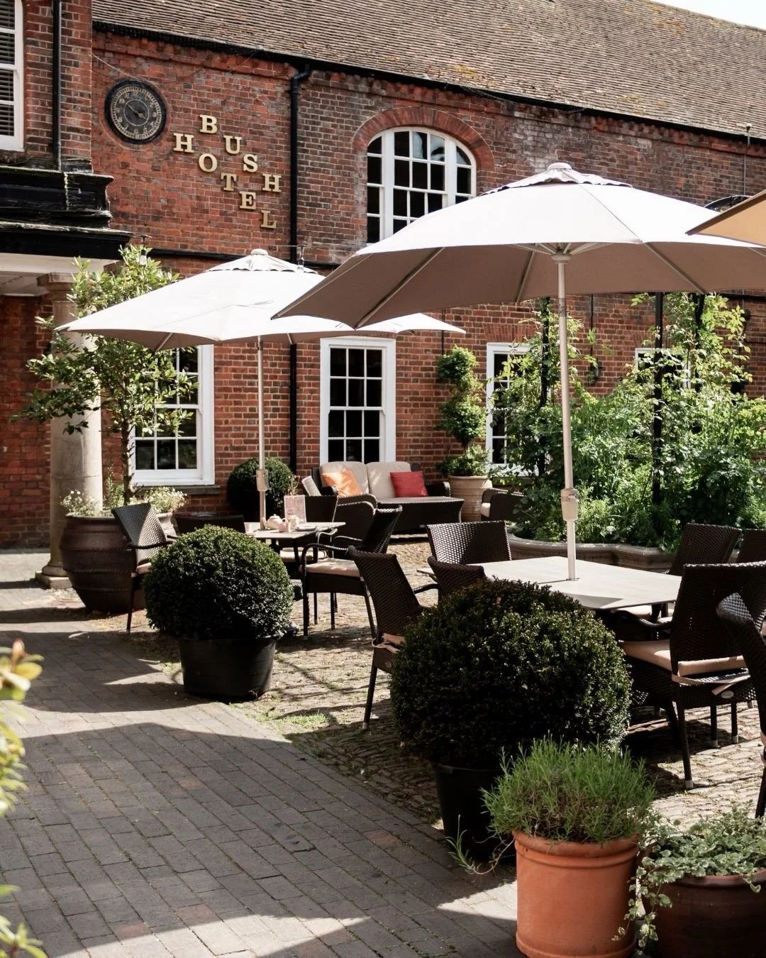 Outdoor patio seating area with tables, chairs, umbrellas, and potted plants in front of a brick building labeled 'BUSH HOTEL'.