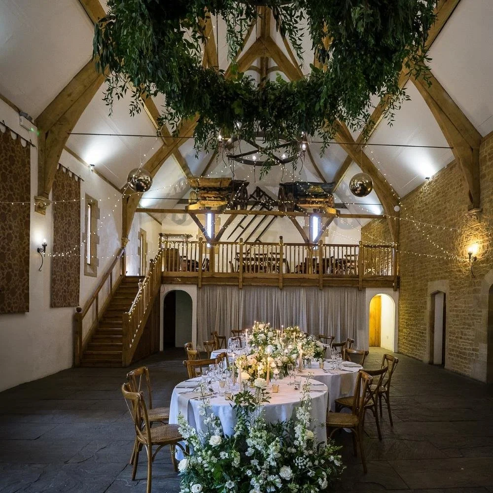 Wedding reception setup with a round table decorated with white flowers, candles, and glassware in a rustic barn with wooden beams, fairy lights, and a second-floor balcony.