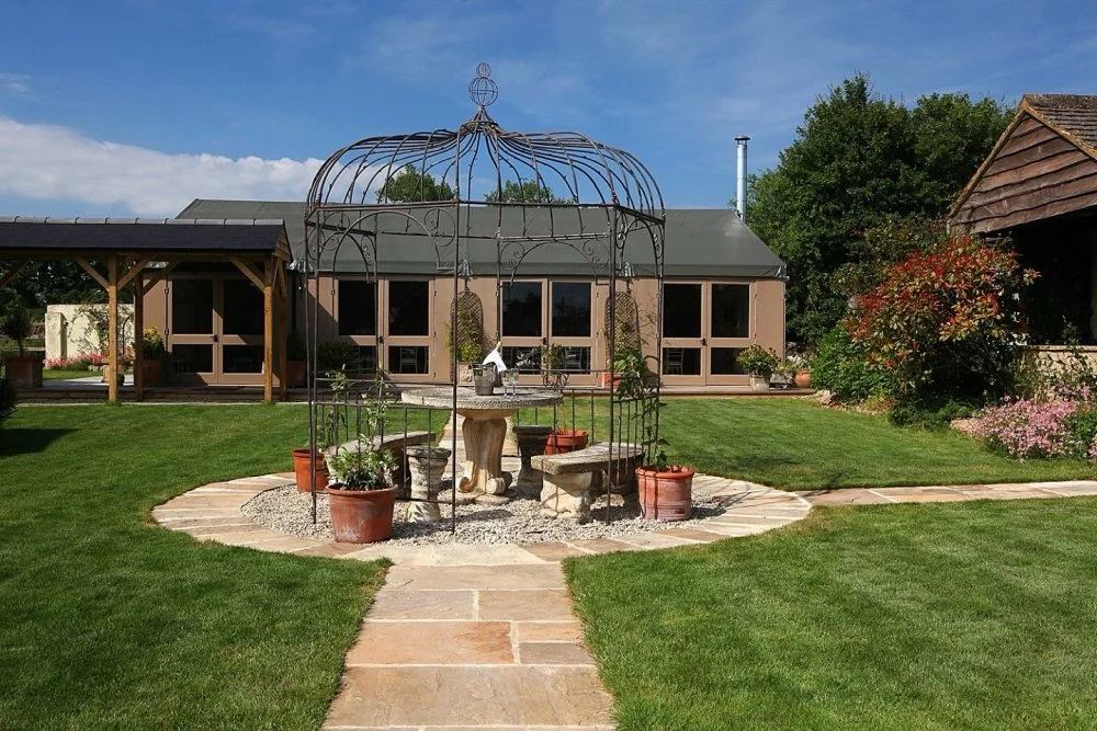 Manicured backyard with stone pathway, decorative metal gazebo, stone table and benches, potted plants, lush green lawn, garden shed, trees, and flowering bushes under a blue sky.