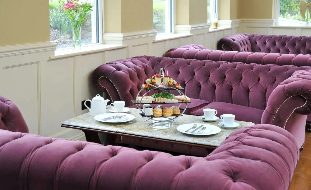 A breakfast setting with a marble table surrounded by purple tufted sofas. On the table is a teapot, cups, plates, silverware, and a three-tiered stand with sandwiches, pastries, and fruit. There is a window with green plants outside.