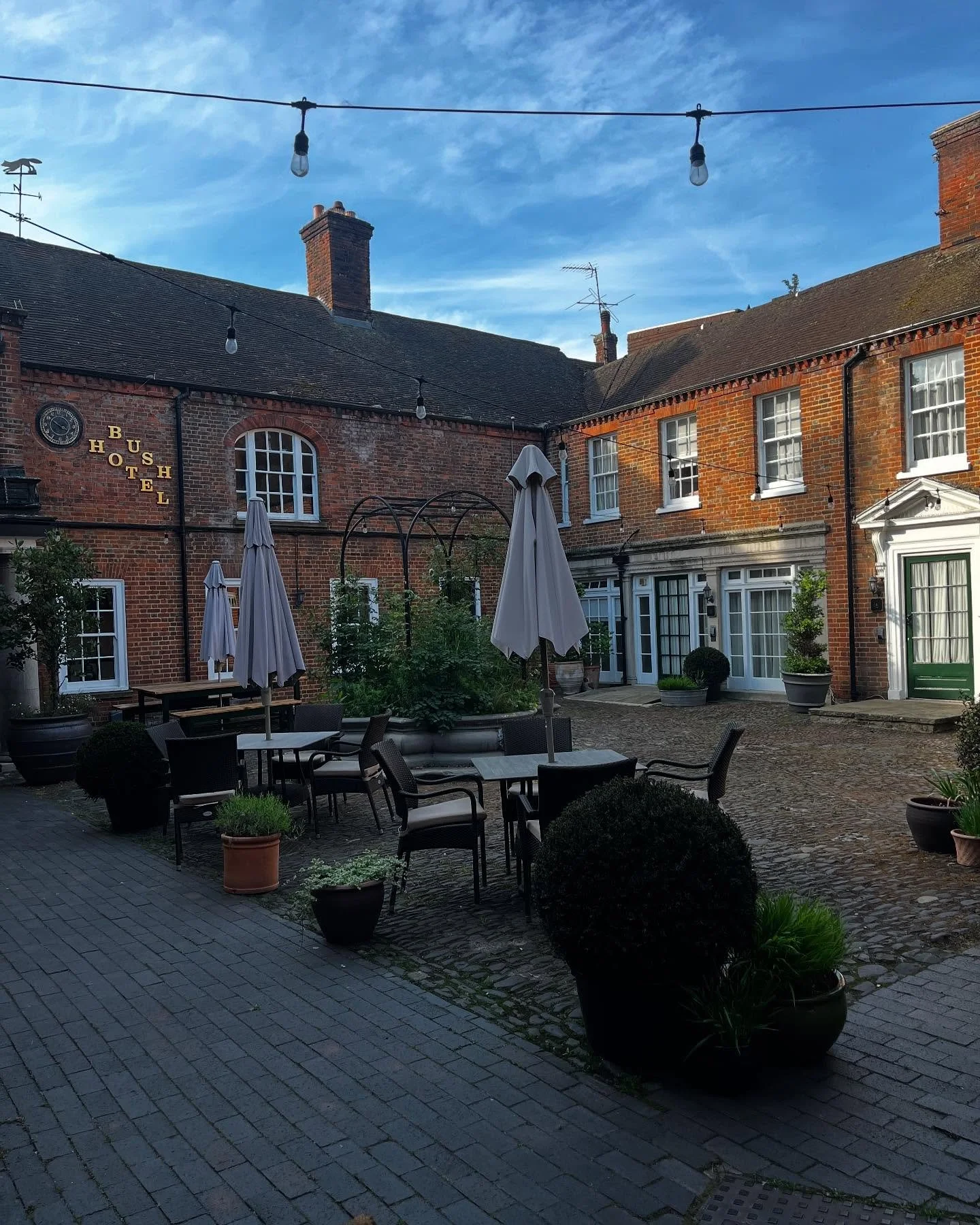 A courtyard with outdoor seating, umbrellas, potted plants, and brick buildings, some with windows and signs, under a partly cloudy sky.