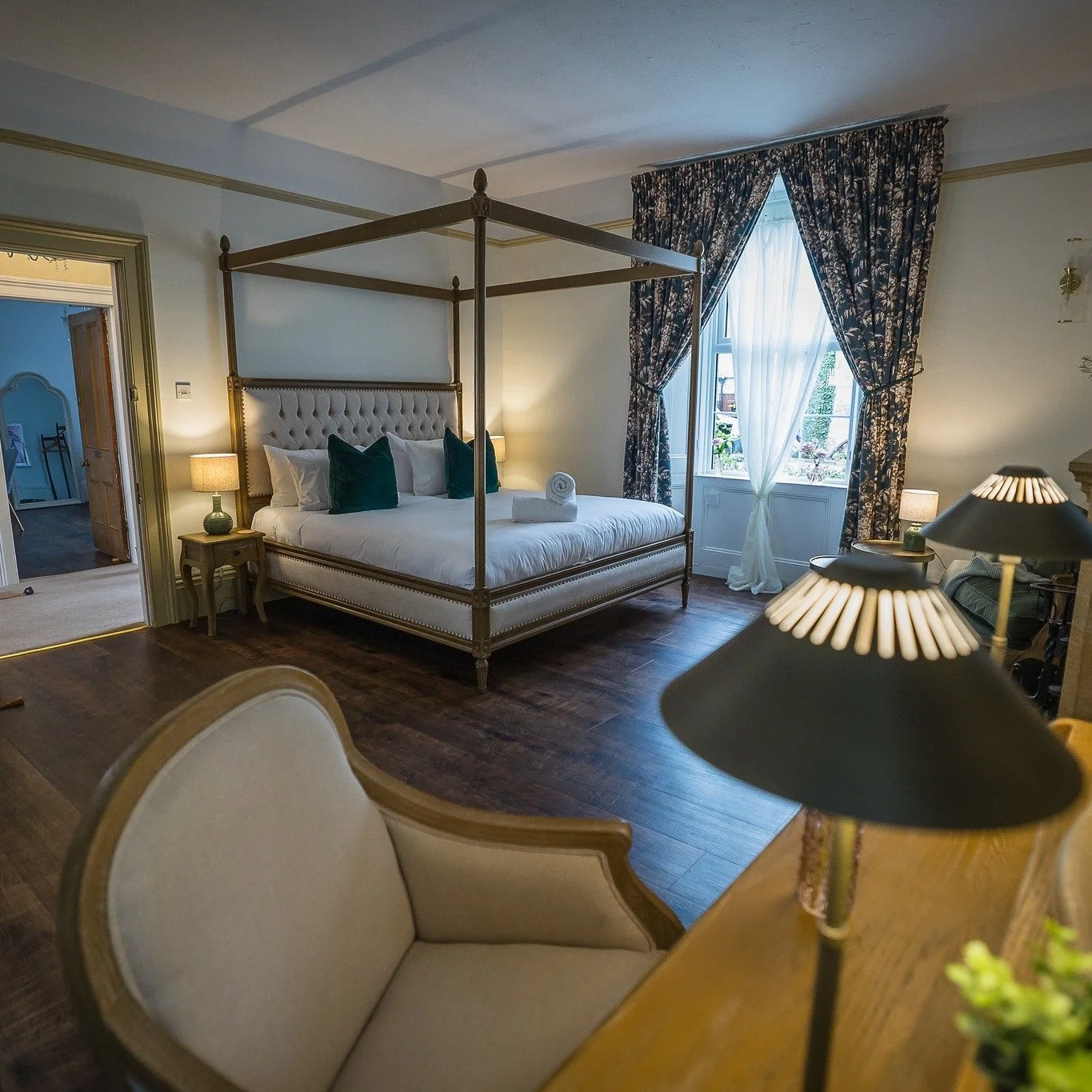 A bedroom with a four-poster bed, bedside lamps, a window with floral curtains, and a cream armchair.