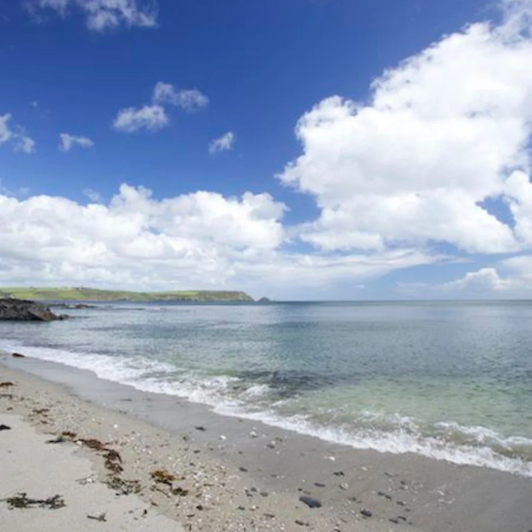 A sandy beach with rocks and seaweed, calm ocean waves, green cliffs in the distance under a blue sky with scattered white clouds.