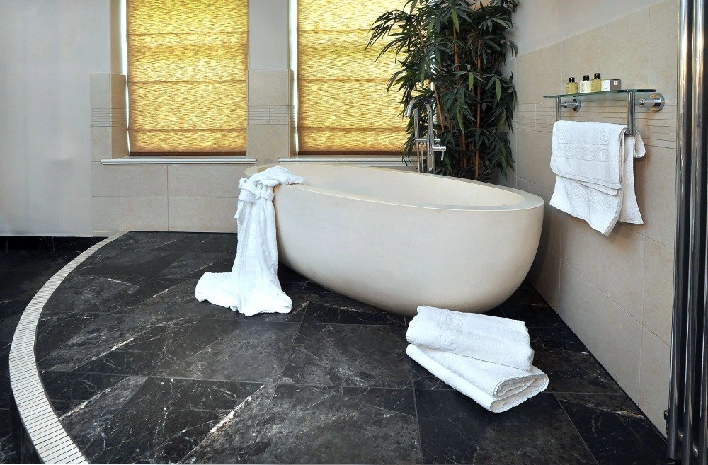 A modern bathroom with a freestanding oval bathtub, white towels, a large green plant, and windows with yellow blinds.